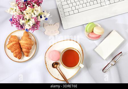 Modern feminine desk, home office. Computer, hyacinth flowers and a cup of tea, macaroons,croissants on a sunny table with long Stock Photo