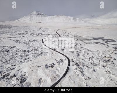 Aerial view of a winding road cuts through the stark white, snowy landscape towards a distant, snow-capped mountain, Budir, Snaefellsbaer, Iceland. Stock Photo