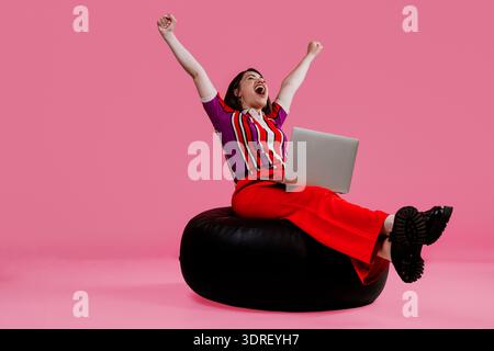 Woman raising arms in excitement, celebrating achievement and triumph while looking at a laptop, sitting on a black bean bag with a vibrant pink backg Stock Photo
