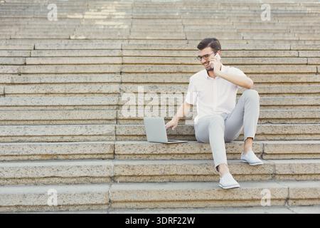 Young Man Works on Laptop While Talking on Phone on Outdoor Steps in Daytime Sunny Weather Stock Photo