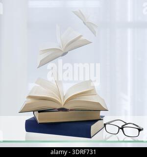 Time to read your favorite books. Old books and black glasses on a glass shelf against open window with transparent curtain and flying books. Lght bac Stock Photo