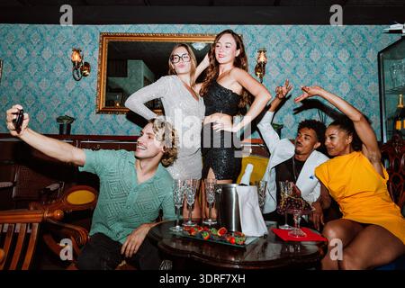 Diverse group of young adult friends smiling and taking a selfie at a party, celebrating a special occasion with champagne and snacks in a stylish bar Stock Photo