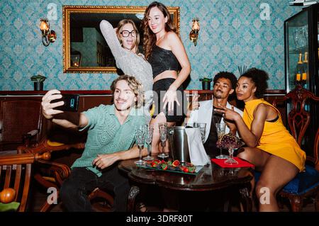 Diverse group of young adult friends smiling and taking a selfie at a party, celebrating a special occasion with champagne and snacks in a stylish bar Stock Photo