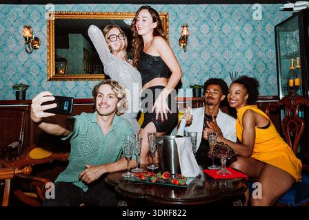 Diverse group of young adult friends smiling and taking a selfie at a party, celebrating a special occasion with champagne and snacks in a stylish bar Stock Photo