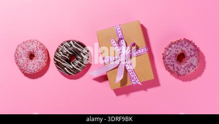 A festive flat lay featuring vibrant donuts with colorful toppings and a gift box on a pink background Stock Photo