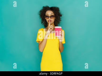 Young Woman in Yellow Dress Holds Coffee Cup and Makes Shushing Gesture Against Blue Background Stock Photo