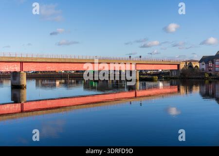 A long red bridge extends over the tranquil River Ness in Inverness, Scotland. Its vibrant color reflects perfectly on the smooth water surface under Stock Photo