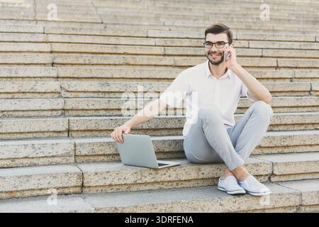 Young Man Sits on Stairs Using Laptop While Talking on Phone During Sunny Day Stock Photo