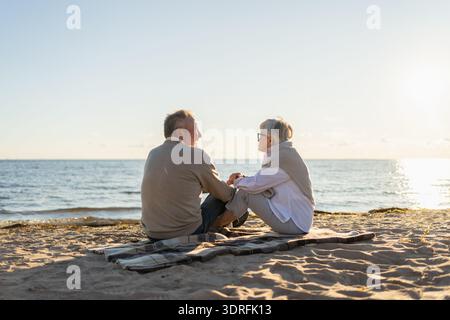 Senior mature couple sitting on beach holding hands enjoying outdoor recreation. Old husband wife touching hands with tenderness Stock Photo