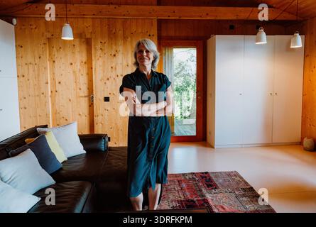 Elegant senior woman with gray hair smiling confidently, arms crossed while standing in a bright, modern living room with pine walls and a cozy couch Stock Photo