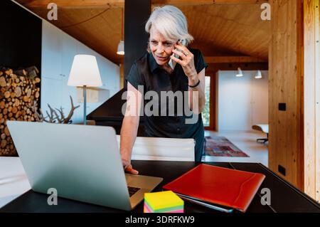 Mature woman leaning on a desk in a modern home office, telecommuting while juggling laptop work and a phone call, focused and confident in a contempo Stock Photo