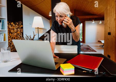 Mature woman leaning on a desk in a modern home office, telecommuting while juggling laptop work and a phone call, focused and confident in a contempo Stock Photo