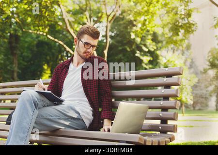 Young Man Sitting on Bench in Park Working on Laptop and Taking Notes Stock Photo