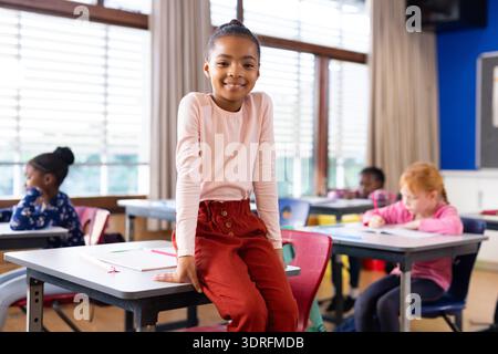 Smiling girl sitting on desk in classroom, students writing in background at school Stock Photo