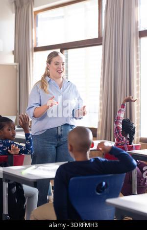 In school, female teacher engaging with diverse students raising hands in classroom discussion Stock Photo