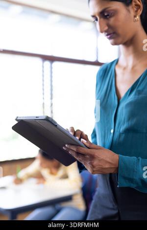 Using tablet, Indian female teacher managing classroom activities while student writing in school Stock Photo