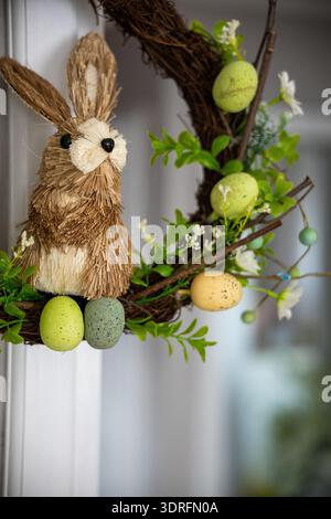 A close-up of a festive Easter wreath hanging on a white door. The charming, handmade decoration features a cute bunny made of straw, colorful speckle Stock Photo