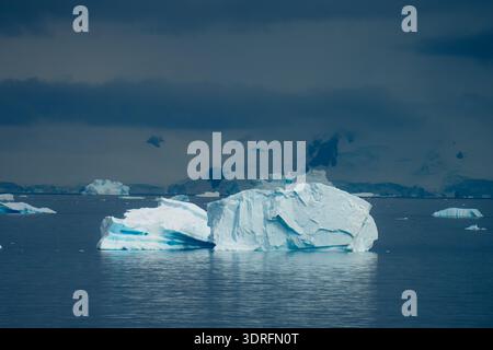 View of a pristine iceberg floats majestically on dark waters under a clouded sky, framed by distant, snow-capped mountains, Portal Point, Antarctica. Stock Photo