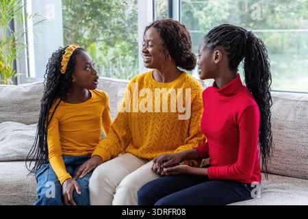 African American mother and daughters talking on gray sofa by large windows holding hands Stock Photo