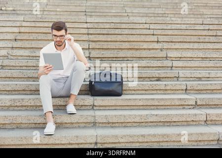 Man Sitting on Steps With Tablet and Bag While Focused on Screen in Urban Area Stock Photo