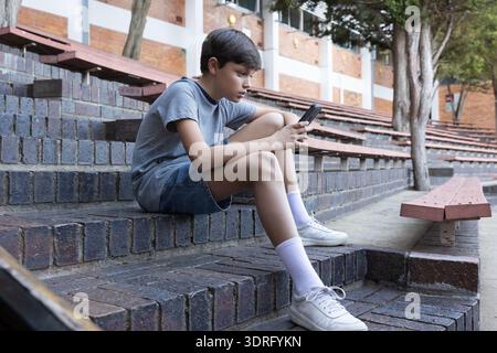 Sitting on steps, boy using smartphone at school, concentrating on screen. Technology, education, concentration, student, connec Stock Photo