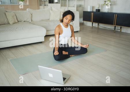 Yoga mindfulness meditation online. African woman practicing yoga with online lessons in laptop at home. Woman sitting in lotus Stock Photo