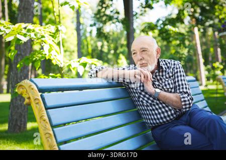 Senior Man Sitting on a Blue Park Bench Looking Thoughtful in a Green Outdoor Area Stock Photo