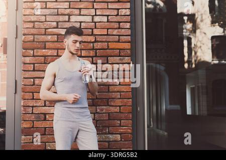 Young Man Stands by Wall Holding a Water Bottle While Resting After Workout Session Stock Photo