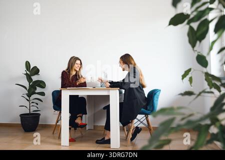 Two women are seated at a table in an office, engaging in conversation and enjoying drinks while discussing work-related topics. Stock Photo