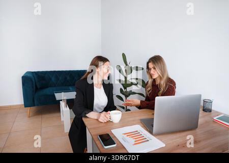 Two women are in an office setting discussing work. One woman is holding a cup while the other gestures at the laptop. Stock Photo