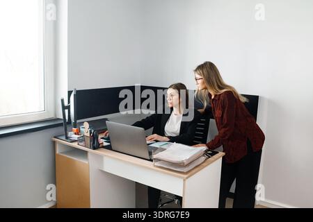 Two women collaborate at a desk in an office setting, focusing on a computer and exchanging ideas during working hours. Stock Photo