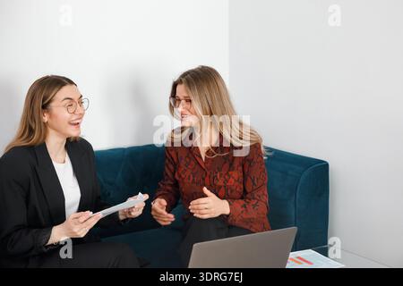 Two women are talking and sharing documents while sitting on a couch in an office. They appear to be collaborating on a task. Stock Photo