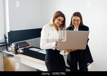 Two women collaborate in an office setting by reviewing content on a laptop while discussing their work tasks. Stock Photo