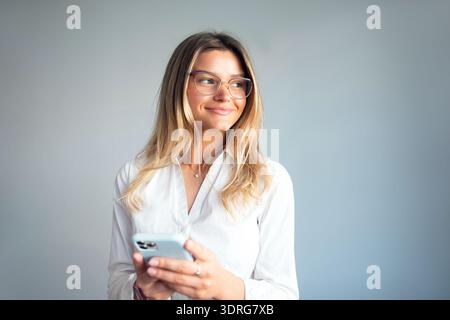 Businesswoman stands in office space, looking at her smartphone with a thoughtful expression while engaged in work activities Stock Photo