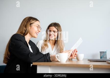 Two women engage in a discussion at a workplace, reviewing documents while enjoying coffee together. Stock Photo