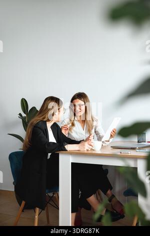 Two women have a conversation at a desk in an office, sharing ideas and discussing projects while sitting in chairs. Stock Photo
