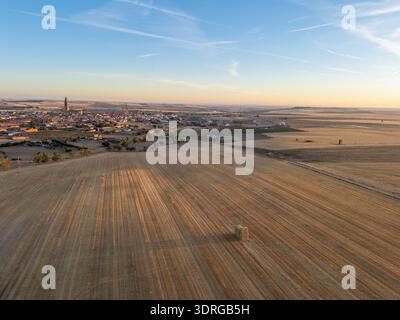 A wide, tranquil rural scene shows harvest fields stretching toward a small village with church towers under a soft sunrise sky. Warm, golden light em Stock Photo