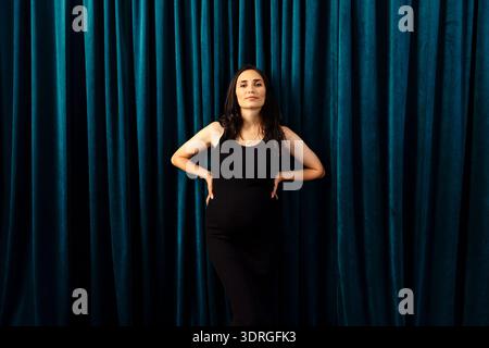 Pregnant woman poses with hands on hips in front of dark green curtains. Charming lady in a black dress looks confidently ahead. Stock Photo