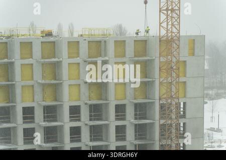 Concrete apartment block framework with repeated balcony modules, insulation covered openings and crane tower, workers installing elements on roof Stock Photo