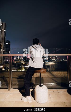 Man sitting on stool at rooftop bar overlooking city skyline at night, urban view with illuminated skyscrapers and glass railing Stock Photo