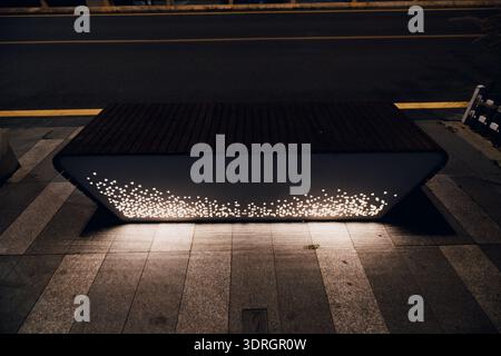 Modern illuminated bench on city sidewalk at night with glowing light dots and urban street background Stock Photo