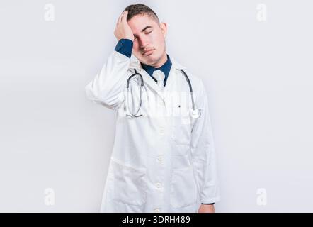Exhausted young doctor with hand on forehead. Tired doctor with hand on forehead isolated Stock Photo