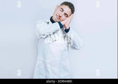 Young doctor making sleep gesture isolated. Handsome doctor making sleep gesture with palms on his cheeks Stock Photo