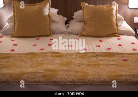 Romantic hotel bed decorated with petals and golden pillows, soft bedside lamps casting warm glow, crisp linen and patterned throw, intimate turndown Stock Photo