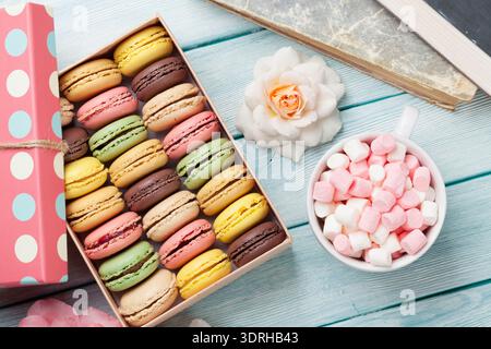 Colorful macaroons in a gift box and marshmallow in coffee cup on wooden table. Sweet macarons and flowers. Top view Stock Photo