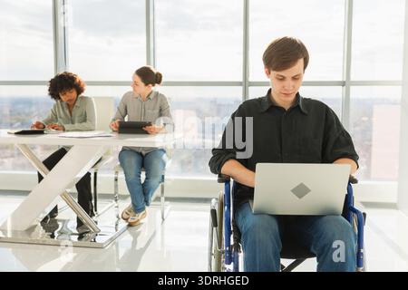 Accessible environment. Young man in chair for people with disability working with laptop in modern office. Diverse multiracial Stock Photo