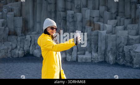 An individual stands on the black sands of Reynisfjara Beach, wearing a bright yellow coat and taking a selfie against towering Stock Photo