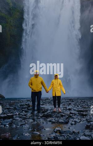 A joyful couple stands hand in hand in front of Skogafoss waterfall in Iceland, wearing bright yellow raincoats. Stock Photo