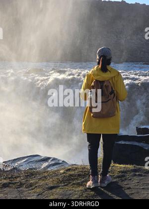 A traveler stands near the majestic Dettifoss waterfall in Iceland, captivated by the roaring cascades and mist rising into the Stock Photo