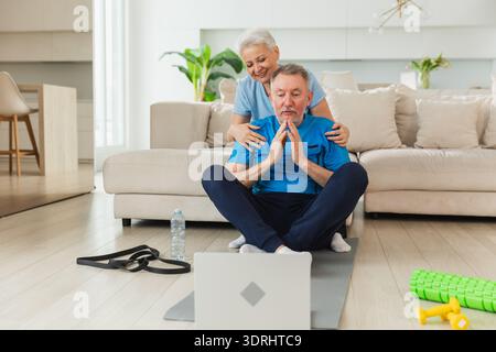 Yoga mindfulness meditation. Senior adult mature couple practicing yoga with online lessons in laptop. Old wife helping teaching Stock Photo